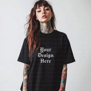 Op de afbeelding: Zwart T-shirt met de tekst "Your Design Here" in een gotisch lettertype. Het shirt heeft korte mouwen en een ronde hals. Het model heeft tatoeages op armen en nek.