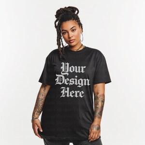 May include: Black t-shirt with the text "Your Design Here" in a gothic font. The shirt is a dark color and has a slightly worn appearance. The model is wearing the shirt and has tattoos on their arms.