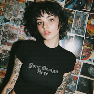 Puede incluir: Top corto negro acanalado con el texto "Your Design Here" en fuente gótica. El top tiene un cuello alto y mangas cortas. El fondo presenta un collage de carteles vintage.