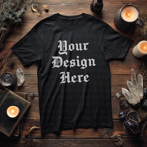 Könnte beinhalten: Schwarzes T-Shirt mit dem Text "Your Design Here" in gotischer Schrift. Das Shirt liegt auf einer Holzoberfläche mit Kerzen, Kristallen und anderen dekorativen Gegenständen, was eine dunkle, mystische Ästhetik erzeugt.