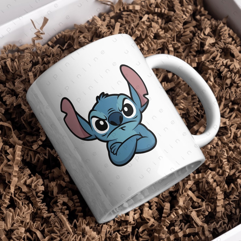 May include: White ceramic mug featuring a blue cartoon character with large ears and a grumpy expression. The character has crossed arms and is set against a white background. The mug has a curved handle and is placed in a box with brown packing material.