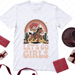 Może przedstawiać: Biała koszulka z kresk&oacute;wkową kowbojką na brązowym koniu, otoczona kaktusami i kapeluszem kowbojskim. Tekst "LET'S GO GIRLS" jest wyświetlany w stylu retro. Zabawna koszulka graficzna o tematyce western.