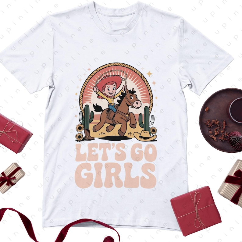Może przedstawiać: Biała koszulka z kresk&oacute;wkową kowbojką na brązowym koniu, otoczona kaktusami i kapeluszem kowbojskim. Tekst "LET'S GO GIRLS" jest wyświetlany w stylu retro. Zabawna koszulka graficzna o tematyce western.