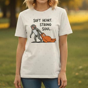 May include: Cream-coloured t-shirt with a cartoon illustration of two monkeys, one grey and one orange, and the words "SOFT HEART. STRONG SOUL." The design is printed on the front of the short-sleeved, round-neck top.