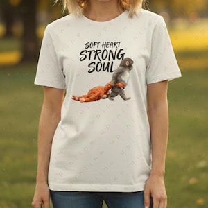 May include: White t-shirt with the text "SOFT HEART STRONG SOUL" in black. The shirt features a graphic of a monkey carrying a stuffed animal. The monkey is grey, and the stuffed animal is orange.