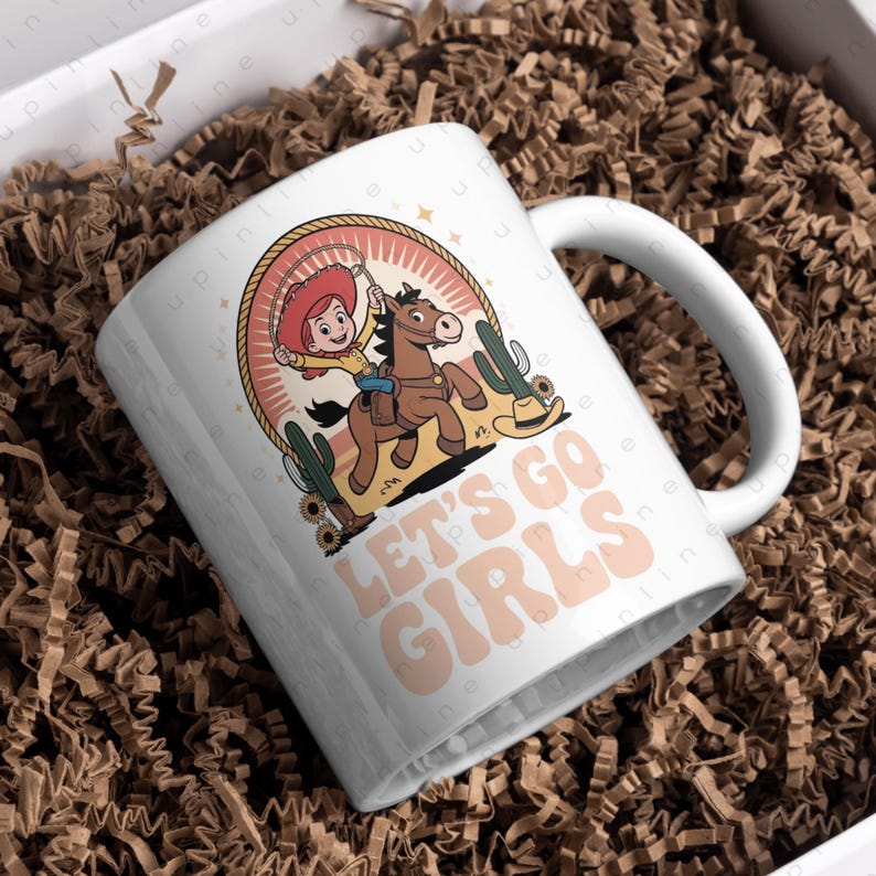 Może przedstawiać: Biały ceramiczny kubek z kresk&oacute;wkową kowbojką na koniu, otoczony kaktusami i słonecznikami. Kubek ma napis "LET'S GO GIRLS" w kolorze brzoskwiniowym. Zapakowany w białe pudełko z brązowym wypełniaczem papierowym.