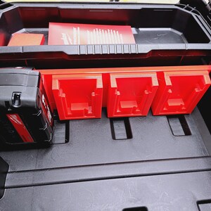 May include: Black plastic tool case with red detailing. The case features a top compartment with a red insert and a lower section with three red storage compartments. A black battery pack is visible.