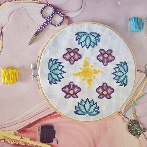 Peut inclure: Un projet de point de croix terminé dans un cercle en bois, avec un motif de soleil jaune central entouré de fleurs violettes et de feuilles turquoise. Les fournitures comprennent des ciseaux, des bobines de fil et un enfile-aiguille, le tout sur une surface rose et dorée.