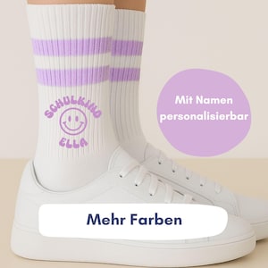Könnte beinhalten: Weiße Sportsocken mit zwei lila Streifen, kombiniert mit weißen Turnschuhen. Die Socken zeigen eine Smiley-Grafik und den Text "SCHULKIND ELLA". Das Bild enthält auch den Text "Mehr Farben".
