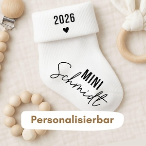 Könnte beinhalten: Weißer Babysocken mit dem Jahr 2026 und einem Herzsymbol, schwarz bedruckt. Der Name "Schmidt" und das Wort "MINI" sind in schwarzer Schrift gedruckt. Das Wort "Personalisierbar" ist in einem weißen Rechteck gedruckt.