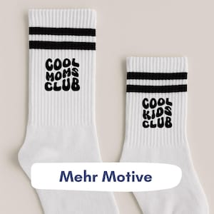 Könnte beinhalten: Weiße Crew-Socken mit schwarzen Streifen am oberen Rand und den Wörtern "COOL MOMS CLUB" und "COOL KIDS CLUB" in Schwarz. Die Socken haben eine gerippte Textur.