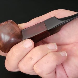 May include: A wooden pipe with a dark brown bowl and a rectangular, reddish-brown stem. The mouthpiece is black and angled. The pipe is held in a hand against a dark background.