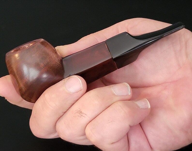 May include: A wooden pipe with a dark brown bowl and a rectangular, reddish-brown stem. The mouthpiece is black and angled. The pipe is held in a hand against a dark background.