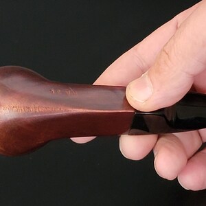 May include: A brown, curved tobacco pipe with a dark, glossy stem. The pipe is held in a hand against a black background. The bowl has a rounded shape and a textured surface.