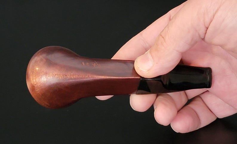 May include: A brown, curved tobacco pipe with a dark, glossy stem. The pipe is held in a hand against a black background. The bowl has a rounded shape and a textured surface.