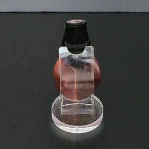 May include: A clear acrylic display stand holding a polished, reddish-brown gemstone. The gemstone is round and has a dark, cylindrical top. The stand has a circular base and a clear, curved support to cradle the stone.