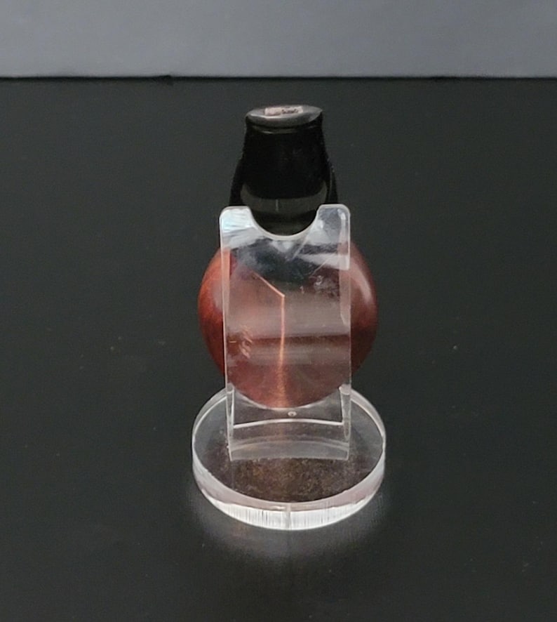 May include: A clear acrylic display stand holding a polished, reddish-brown gemstone. The gemstone is round and has a dark, cylindrical top. The stand has a circular base and a clear, curved support to cradle the stone.