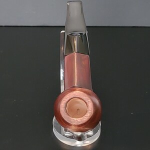 May include: A wooden pipe with a dark brown finish and a clear mouthpiece. The pipe has a rounded bowl and a rectangular stem. It is displayed on a clear acrylic stand.