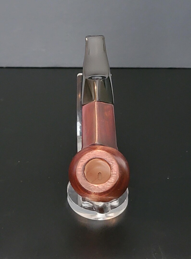 May include: A wooden pipe with a dark brown finish and a clear mouthpiece. The pipe has a rounded bowl and a rectangular stem. It is displayed on a clear acrylic stand.