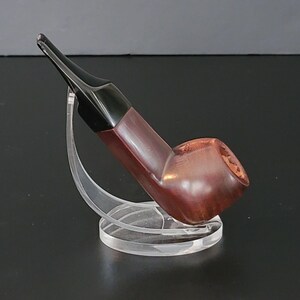 May include: A wooden tobacco pipe with a dark brown bowl and a black stem. The pipe is displayed on a clear acrylic stand, showcasing its curved design and polished finish. The pipe's bowl has a textured surface.