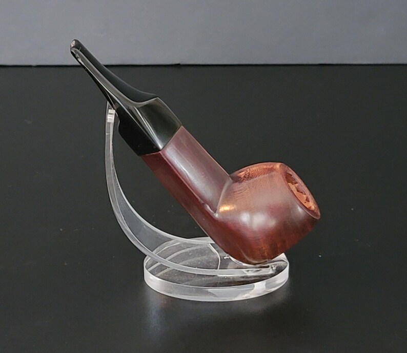 May include: A wooden tobacco pipe with a dark brown bowl and a black stem. The pipe is displayed on a clear acrylic stand, showcasing its curved design and polished finish. The pipe's bowl has a textured surface.