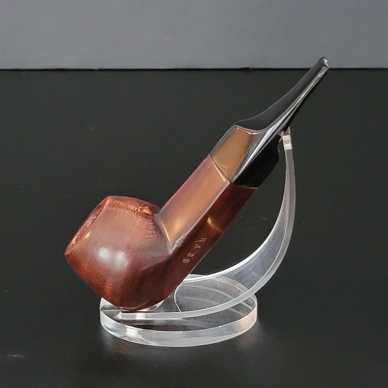 May include: A brown and black tobacco pipe with a curved stem, resting on a clear acrylic stand. The pipe has a rounded bowl and a smooth, polished finish. The stem is black, and the body of the pipe is a rich, dark brown color. The text "Ra 30" is visible on the pipe.