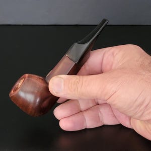 May include: A wooden tobacco pipe with a dark brown bowl and a black stem. The pipe is held in a hand against a dark background. The bowl is rounded and polished, and the stem is angled.