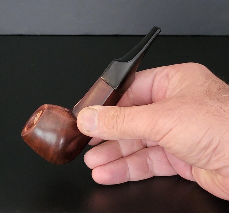 May include: A wooden tobacco pipe with a dark brown bowl and a black stem. The pipe is held in a hand against a dark background. The bowl is rounded and polished, and the stem is angled.