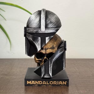 May include: A black and silver Mandalorian helmet bust with a gold skull, mounted on a black base. The base has the word "MANDALORIAN" in gold lettering. The bust is a detailed representation of the character's helmet and armor.