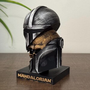 May include: A Mandalorian helmet bust in black and silver, with a gold-colored skull detail. The bust sits on a black base with the word "MANDALORIAN" in gold lettering. The helmet has a weathered, metallic appearance.