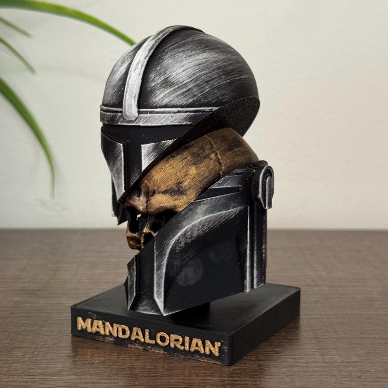 May include: A Mandalorian helmet bust in black and silver, with a gold-colored skull detail. The bust sits on a black base with the word "MANDALORIAN" in gold lettering. The helmet has a weathered, metallic appearance.