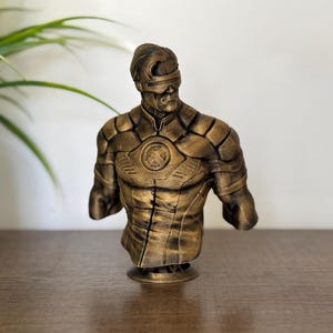 May include: A gold-colored bust of a superhero figure with a visor and a stylized 'X' on the chest. The figure is standing on a small round base.