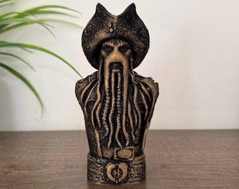 May include: A detailed bust of Davy Jones, a character from the Pirates of the Caribbean film series. The sculpture features a dark, weathered finish with gold accents. The bust includes Jones' iconic hat, beard, and tentacle-like facial features.