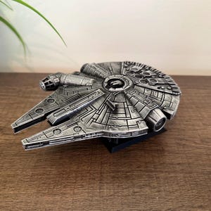 May include: A detailed, silver-colored model of the Millennium Falcon starship. The model features intricate paneling and design elements, with a dark base. The ship's iconic shape is clearly visible, showcasing its unique features.