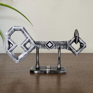 May include: A silver-colored key with intricate geometric designs, displayed on a matching stand. The key has a large, octagonal head and a long, narrow shaft with repeating patterns.