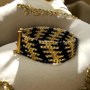 Puede incluir: Una pulsera de cuentas negras y doradas con un diseño geométrico. La pulsera está hecha de pequeñas cuentas y tiene cierres de metal dorado. También se ven una cadena de oro y otras joyas de oro.