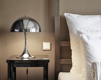 May include: A chrome table lamp with a rounded shade and a slender stem sits on a black nightstand. The lamp reflects the room's surroundings. The nightstand is next to a bed with white pillows and a wooden headboard. The wall is a neutral brown.