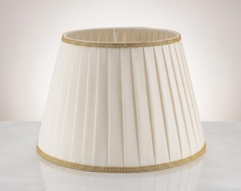 Puede incluir: Una pantalla de lámpara en forma de tambor de color crema, con pliegues verticales en todo el exterior. Los bordes superior e inferior están rematados con un ribete decorativo texturizado de color dorado que enmarca la tela plisada.