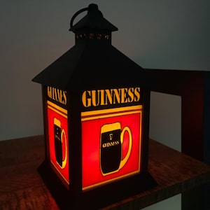 Peut inclure: Lanterne en métal noir avec le logo Guinness et un verre de Guinness sur fond rouge. La lanterne est éclairée, avec le mot "GUINNESS" en lettres dorées. Un objet décoratif pour un pub ou un bar à domicile, avec une poignée supérieure.