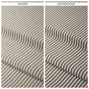 Puede incluir: Dos muestras de tela con rayas paralelas onduladas. La muestra de la izquierda está etiquetada como "VELINOR" y presenta rayas marrones claras y blancas. La muestra de la derecha, etiquetada como "AUTUMN BLAZE", tiene rayas marrones y blancas.