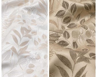 Könnte beinhalten: Zwei Stoffmuster, eines mit der Aufschrift "WISTFUL" und das andere mit der Aufschrift "RAIN SAND". Der Stoff "WISTFUL" ist cremefarben mit einem dezenten Blattmuster in Beige. Der Stoff "RAIN SAND" ist beige mit einem dunkleren beigen Blattmuster. Beide Stoffe haben einen Satin-Glanz.