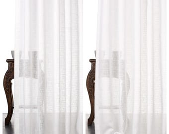 Puede incluir: Dos cortinas de lino blanco, etiquetadas como "WHITE MIRAGE" y "SWEET CHIVES", cuelgan frente a una mesa de madera oscura. Las cortinas son ligeramente translúcidas, permitiendo el paso de algo de luz. La tela tiene una apariencia texturizada y las cortinas llegan hasta el suelo.