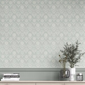 Op de afbeelding: Lichtgroen behang met een herhalend wit bloemenpatroon. De muur is versierd met een decoratieve lijst en een houten plank. Een vaas met planten en boeken staat op de plank.