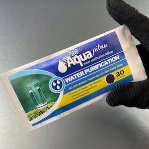 Puede incluir: Un paquete de tabletas de purificación de agua Aqua Pitna, sostenido por una mano enguantada. El paquete es blanco, azul y amarillo, con texto que indica que es para la purificación del agua y contiene 30 tabletas. La imagen incluye un gráfico de un vaso de agua.