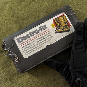 May include: A gray leather wallet with the text "Electro-fix" and a list of tools printed on a label. The wallet has a zipper and a small lightbulb charm attached. A black glove is partially visible.