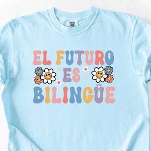 Pu&ograve; includere: Una maglietta azzurra con la scritta "EL FUTURO ES BILINGUE" in lettere colorate in stile retr&ograve;. Il design include grafiche floreali. La maglietta &egrave; realizzata in materiale morbido ed &egrave; piegata su una superficie bianca.
