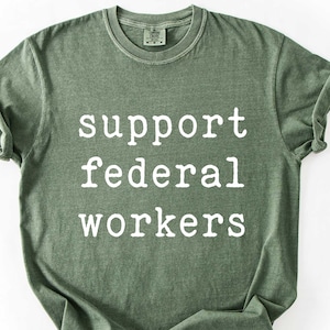 May include: A moss green t-shirt with the words "support federal workers" printed in white. The shirt has a crew neck and rolled-up sleeves. The word "MOSS" is in a small orange rectangle in the bottom right corner.