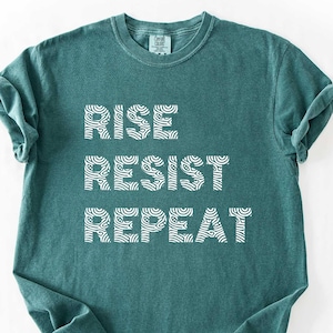 May include: A teal-colored t-shirt with the words "RISE RESIST REPEAT" printed in white. The shirt has rolled-up sleeves and a crew neck. The bottom right corner of the image has a rectangular label that says "BLUE SPRUCE".