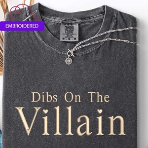 May include: A dark gray t-shirt with the phrase "Dibs On The Villain" in a gold-colored font. The shirt is styled with a silver necklace and a pair of blue jeans. The word "Embroidered" is on a purple tag.