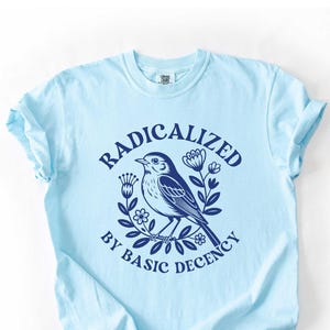 May include: Light blue t-shirt with a navy blue graphic of a bird surrounded by flowers and the text "RADICALIZED BY BASIC DECENCY". The shirt is made of soft material and has rolled-up sleeves.